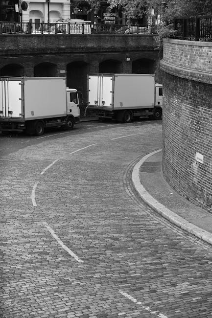 Black and white photograph of a narrow cobblestone street with a curved layout, showing three boxes trucks parked along the side just outside a brick underpass and near a brick wall. The trucks are positioned parallel to the curb, with the rear doors facing the viewer. In the background, there is a bridge with a railing and some trees, indicating an urban environment. The scene captures the loading process typical of house removals, where furniture and packing materials would be loaded onto the delivery vehicles. The cobblestone surface and brick structures suggest the setting is in a historic or older part of Lewisham. Man and Van Lewisham’s removal services may utilise such a street for furniture transport and home relocation logistics, involving loading and unloading items into the vans amidst the street environment. The overall composition highlights vehicle positioning and street layout relevant to professional removals.