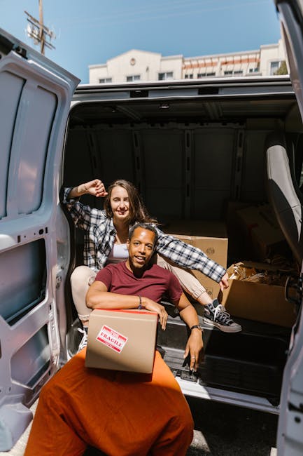Two smiling women are engaged in a home relocation process, with one seated on a box marked 'fragile' inside the open rear of a light grey van and the other standing behind them, holding a small cardboard box. The van is parked outside a residential building with multiple stories visible in the background, under a blue sky. Inside the van, visible items include various cardboard boxes, some wrapped in plastic or fabric for protection, and a wooden furniture piece. The loading area appears organized, with the women actively involved in packing or preparing furniture and boxes for transport. The scene captures the typical process of furniture transport and packing during a house move, with equipment such as boxes, protective wrapping, and the van ready for the next stage of the move, reflecting professional removal service practices by Man and Van Lewisham.