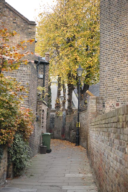 A narrow, paved alleyway between brick buildings with a tall tree bearing yellow autumn leaves at the far end. On the left side, there are bushes with orange and green foliage, a green wheelie bin, and a small black trash bin. On the right, a high brick wall extends along the length of the alley. An old-fashioned black lamppost with a yellow ribbon tied around it stands near the center, providing ambient outdoor lighting. The area appears quiet and residential, suitable for a house removal or home relocation process. This alleyway, with its compact layout and fallen leaves scattered on the pavement, illustrates a typical scene where furniture and boxes might be carried or transported during a move, supported by a professional removal service such as Man and Van Lewisham, who manage the logistics of moving furniture and household items through tight urban spaces near Blackheath Common, LEWISHAM.