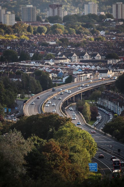 Aerial view of a multi-lane highway with cars and vehicles moving in both directions, curving around a wooded area with dense green trees, leading into an urban residential neighbourhood featuring terraced houses and various buildings. In the background, there are taller apartment blocks and high-rise buildings set against a backdrop of distant hills and greenery, under a partly cloudy sky. The scene captures a typical cityscape with traffic during daylight hours, possibly during a home relocation process involving professional furniture transport and moving logistics, as provided by services like Man and Van Lewisham, who specialise in removals in the Blackheath area near Blackheath Common, LEWISHAM.