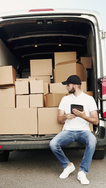 A man wearing a black cap, white t-shirt, and blue jeans is seated on the edge of an open van, which is filled with sealed brown cardboard boxes of various sizes, some stacked on top of each other. The van is parked outdoors on a paved surface, with the back doors open, revealing the interior loaded with moving boxes, likely prepared for home relocation or furniture transport. The man is holding a digital tablet, possibly coordinating packing, loading, or logistics for a moving service. The environment appears well-lit, suggesting daytime, with no other vehicles or objects visible in the immediate surroundings. The scene reflects the packing and loading process involved in professional removals, aligned with the services offered by Man and Van Lewisham, especially as part of a home moving or furniture transport operation, supporting the context of house removals or relocation services relevant to the Catford Broadway area.
