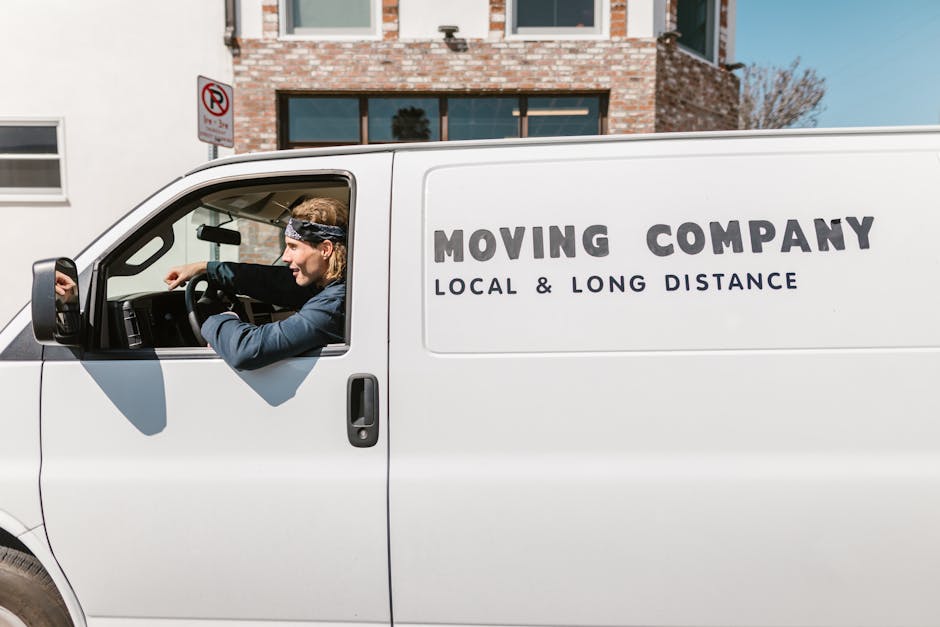 A white moving van with the words 'MOVING COMPANY, LOCAL & LONG DISTANCE' printed on its side is parked on a street in front of a residential building with brick and white facades. Inside the driver's cab, a man with light-colored, shoulder-length hair, wearing a black and white patterned bandana, a blue jacket, and black gloves, is seated and appears to be preparing for loading or driving. The van is positioned on the pavement, with the driver's side door open, and the window rolled down. The scene is illuminated by daylight, and the background shows a tree with bare branches and a 'No Parking' sign mounted on a post near the building. This setting suggests home relocation or furniture transport as part of a professional removals service provided by Man and Van Lewisham, supporting house moves and moving logistics.