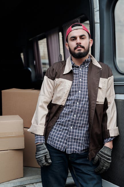 A man wearing a black cap, white t-shirt, and blue jeans is seated on the edge of an open van, which is filled with sealed brown cardboard boxes of various sizes, some stacked on top of each other. The van is parked outdoors on a paved surface, with the back doors open, revealing the interior loaded with moving boxes, likely prepared for home relocation or furniture transport. The man is holding a digital tablet, possibly coordinating packing, loading, or logistics for a moving service. The environment appears well-lit, suggesting daytime, with no other vehicles or objects visible in the immediate surroundings. The scene reflects the packing and loading process involved in professional removals, aligned with the services offered by Man and Van Lewisham, especially as part of a home moving or furniture transport operation, supporting the context of house removals or relocation services relevant to the Catford Broadway area.