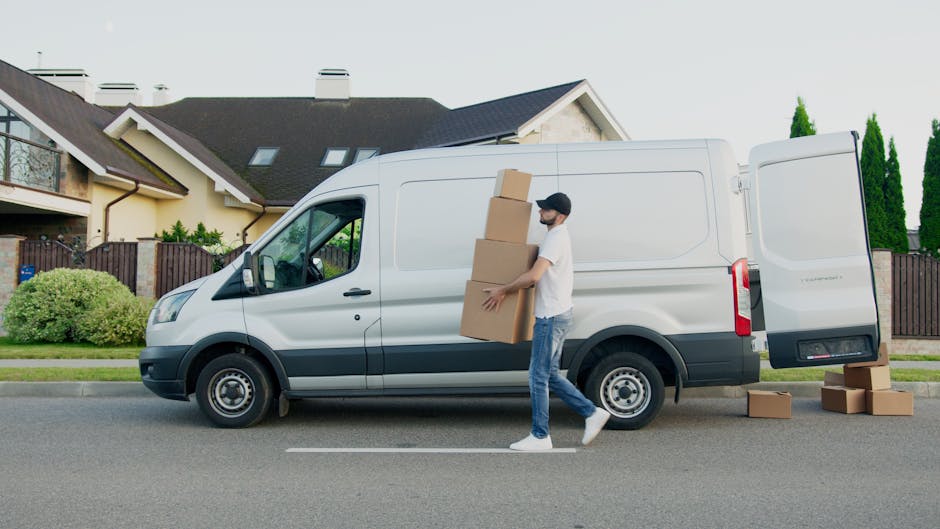 A man wearing a white t-shirt, blue jeans, white sneakers, and a black cap is seen walking alongside a white cargo van with its rear door open. He is carrying three cardboard boxes stacked in his arms