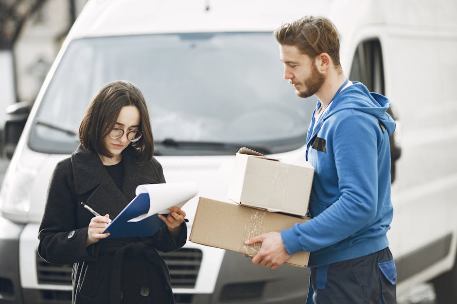 A man in a blue hoodie is holding two cardboard boxes, one stacked on top of the other, and appears to be loading or unloading them near a white van, likely during a home relocation or furniture transport process. A woman in glasses and a dark coat is standing nearby, taking notes or checking a clipboard, possibly coordinating the move or managing packing details, inside a parking area or driveway. The background features the front of a white commercial van with a slightly blurred urban environment, including other vehicles and buildings, with natural daylight illuminating the scene. This image illustrates a professional removal service, with emphasis on packing, moving logistics, and transportation activities relevant to house removals or moving services, as provided by companies like Man and Van Lewisham.