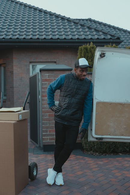 A man standing outside a residential property near a white van engaged in the home relocation process. He is dressed in a blue long-sleeve shirt, black trousers, a black padded vest, and white footwear, with one hand resting on his hip. The van, positioned on the driveway, has its rear door open, revealing an interior likely used for transporting furniture and boxes. In the foreground, there are cardboard boxes and packaging materials, indicating packing and moving activities. The suburban house features a brick wall, a window, and a tiled roof, with a small garden area visible behind the van. The scene captures the loading process typical of professional removals, supporting seamless furniture transport and household relocation services offered by Man and Van Lewisham.