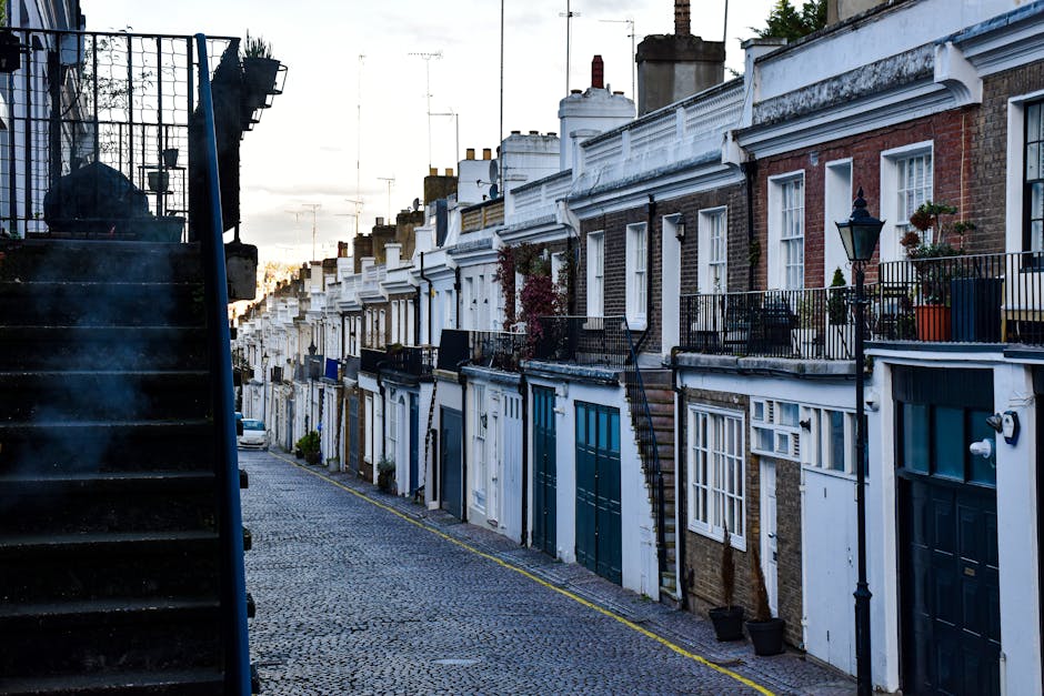 A narrow cobbled street in Lewisham with a row of terraced homes featuring brick and white-rendered facades, some with small front gardens and potted plants. The buildings have upper-level balconies with black metal railings, and one property has a black staircase leading to a side entrance. To the left, a black metal staircase with a diamond-patterned railing extends upward, partially visible in the foreground. The street is illuminated by soft natural light, likely during early evening or late afternoon, with several chimneys visible atop the rooftops. The scene depicts a quiet residential area, possibly a challenging location for furniture transport during home relocation, where careful planning and specialist removals services like those from Man and Van Lewisham are often required due to the steep streets and narrow passageways.