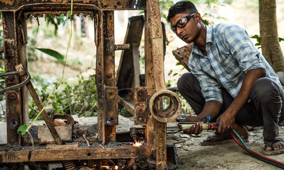 A man with dark hair, glasses, and wearing a blue and white checked shirt while crouching outdoors next to a rusty, partially disassembled metal structure, uses a handheld welding or cutting tool to work on the metal frame. The scene is set in a natural environment with green foliage and tree trunks visible in the background, indicating an outdoor workspace. The man is focused on his task, and the equipment and metal materials suggest a process of repair or modification, which may relate to structural or equipment transportation preparations during home relocations or furniture transport. This detailed view highlights the physical effort involved in moving-related tasks such as dismantling or restoring large items, and occasional use of tools like welders or cutters fits into the context of thorough packing, disassembly, and logistical management by services like Man and Van Lewisham.