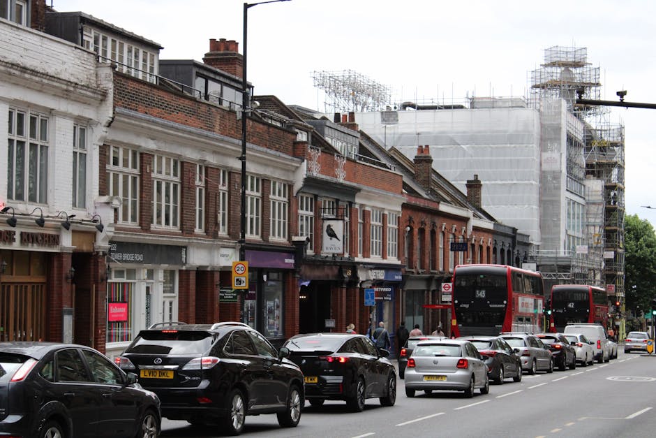 A narrow cobbled street in Lewisham with a row of terraced homes featuring brick and white-rendered facades, some with small front gardens and potted plants. The buildings have upper-level balconies with black metal railings, and one property has a black staircase leading to a side entrance. To the left, a black metal staircase with a diamond-patterned railing extends upward, partially visible in the foreground. The street is illuminated by soft natural light, likely during early evening or late afternoon, with several chimneys visible atop the rooftops. The scene depicts a quiet residential area, possibly a challenging location for furniture transport during home relocation, where careful planning and specialist removals services like those from Man and Van Lewisham are often required due to the steep streets and narrow passageways.