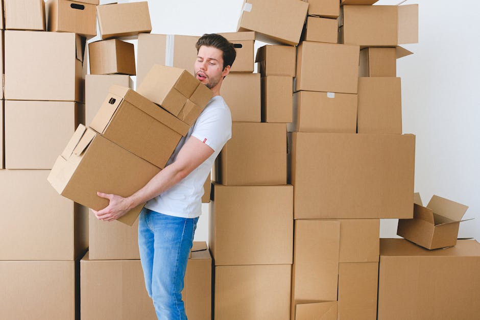 A man with dark hair and a light beard is carrying a medium-sized cardboard box, carefully holding it with both hands, in front of a large stack of similarly packed cardboard boxes of various sizes. The boxes are arranged in a disorderly manner, some stacked on top of each other, others leaning against the white wall of an interior room. The man is dressed in a white t-shirt and blue jeans, and appears focused on moving the box safely. The scene is well-lit, highlighting the materials used for packing, such as cardboard and paper, which are typical for a home relocation or moving process. This image illustrates the logistics of packing and furniture transport as part of a house removal, with a focus on the careful handling of items during an internal packing and moving task, associated with professional services like those offered by Man and Van Lewisham.