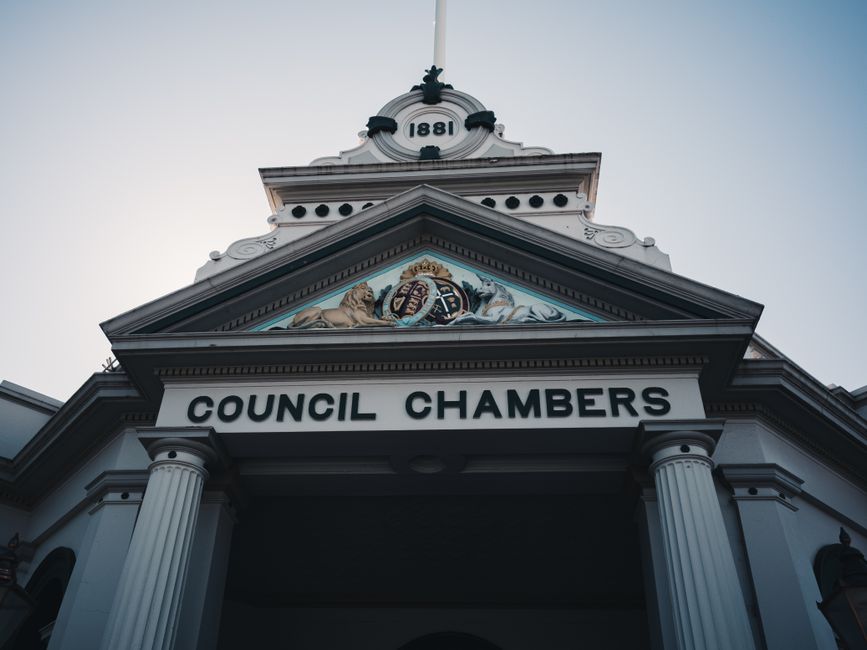 Exterior view of a historic council chambers building with a white façade and decorative architectural details, including columns and a triangular pediment featuring a crest and the year '1881' at the top, captured in daylight with a clear sky background. The building serves as a key local government venue in Lewisham, relevant to residents considering council permit charges for house and furniture removals. The image illustrates the formal entrance area, emphasizing the building's traditional style, which may be associated with procedures involved in home relocation and the need for appropriate permits. The presence of structural elements such as columns and classic decorative motifs aligns subtly with the logistics of packing, loading, and transporting furniture during a removal process, underscoring the importance of adhering to local regulations when planning a move within Lewisham. As part of Lewisham removals, [COMPANY_NAME] understands these procedures and the importance of proper planning for efficient furniture transport.