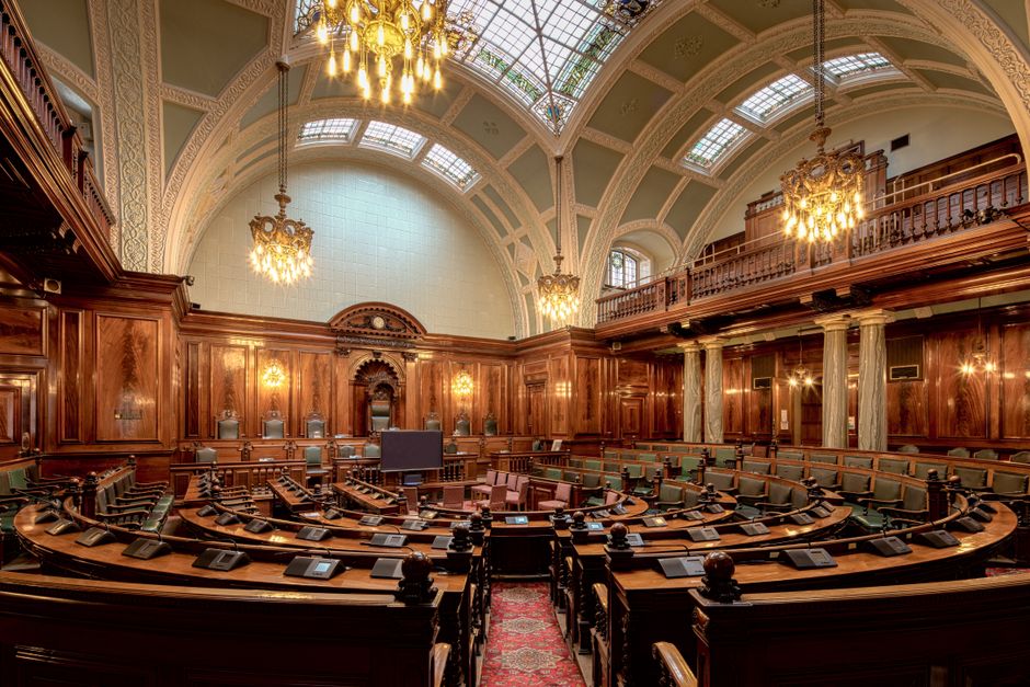 A spacious and ornately decorated parliamentary chamber featuring a central semicircular seating arrangement with multiple rows of green upholstered benches and dark wooden desks, each equipped with a black microphone and electronic voting device. The room has high, arched ceilings with intricate architectural details and large stained glass windows allowing natural light to illuminate the space. Chandeliers hang from the ceiling, casting warm lighting across the wooden-paneled walls and upper gallery, which is supported by marble columns. The chamber is currently empty, with no people present, and the focus is on the detailed woodwork, lighting fixtures, and overall grandeur of the interior as seen in a professional setting related to government or legislative activities, suitable for context about house removals or relocation involving sensitive sites like parliament buildings.